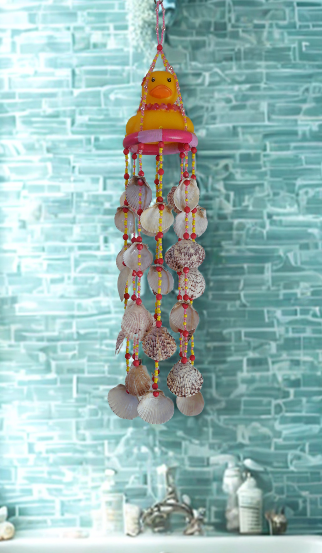 Decorative shell wind chime with a rubber duck hanging against a light blue tiled wall.