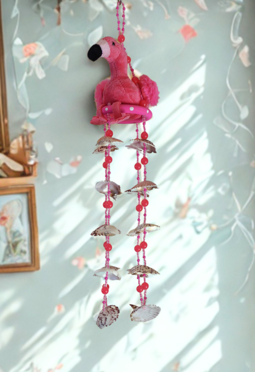 Pink flamingo wind chime with shells hanging against a light blue wall.
