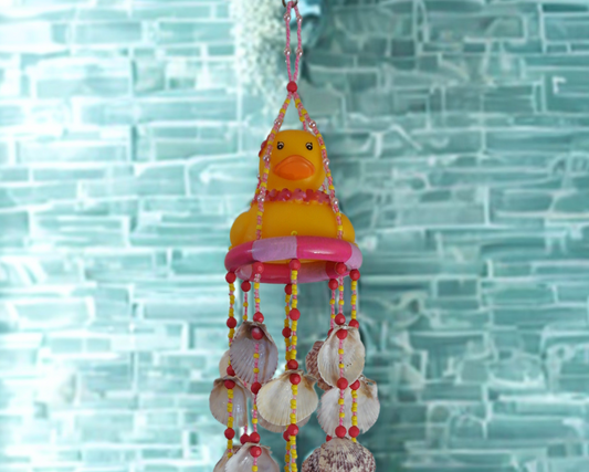 Decorative wind chime with a yellow rubber duck and shell beads against a light blue brick wall.